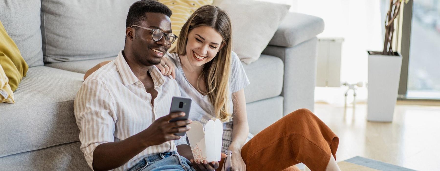 a man and woman with take out, sit against a couch on their living room floor and watch at their cell phone